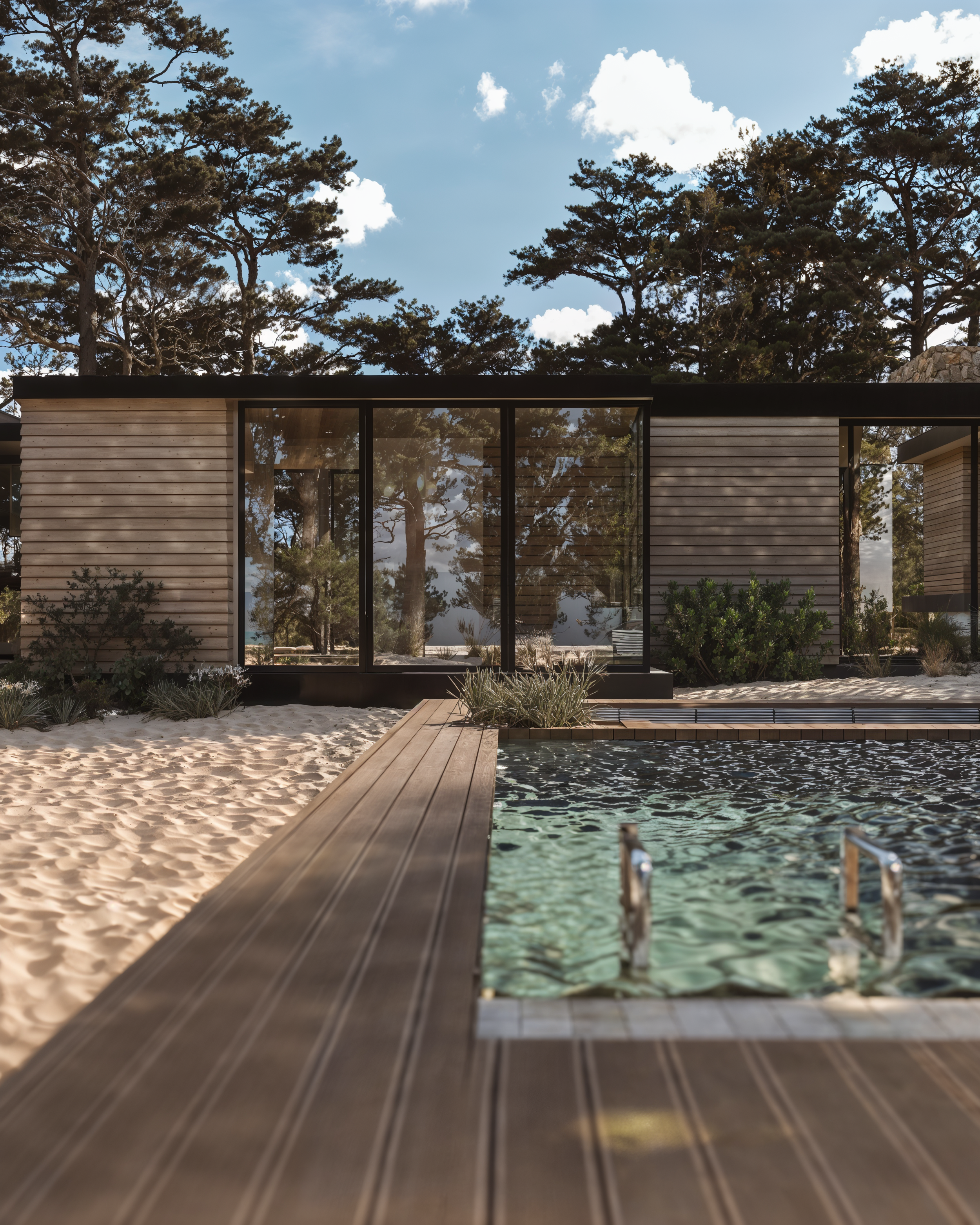 Casa moderna de vidro com paredes de janelas grandes, situada ao ar livre, rodeada por árvores e vegetação, com piscina em primeiro plano e céu com nuvens ao fundo.