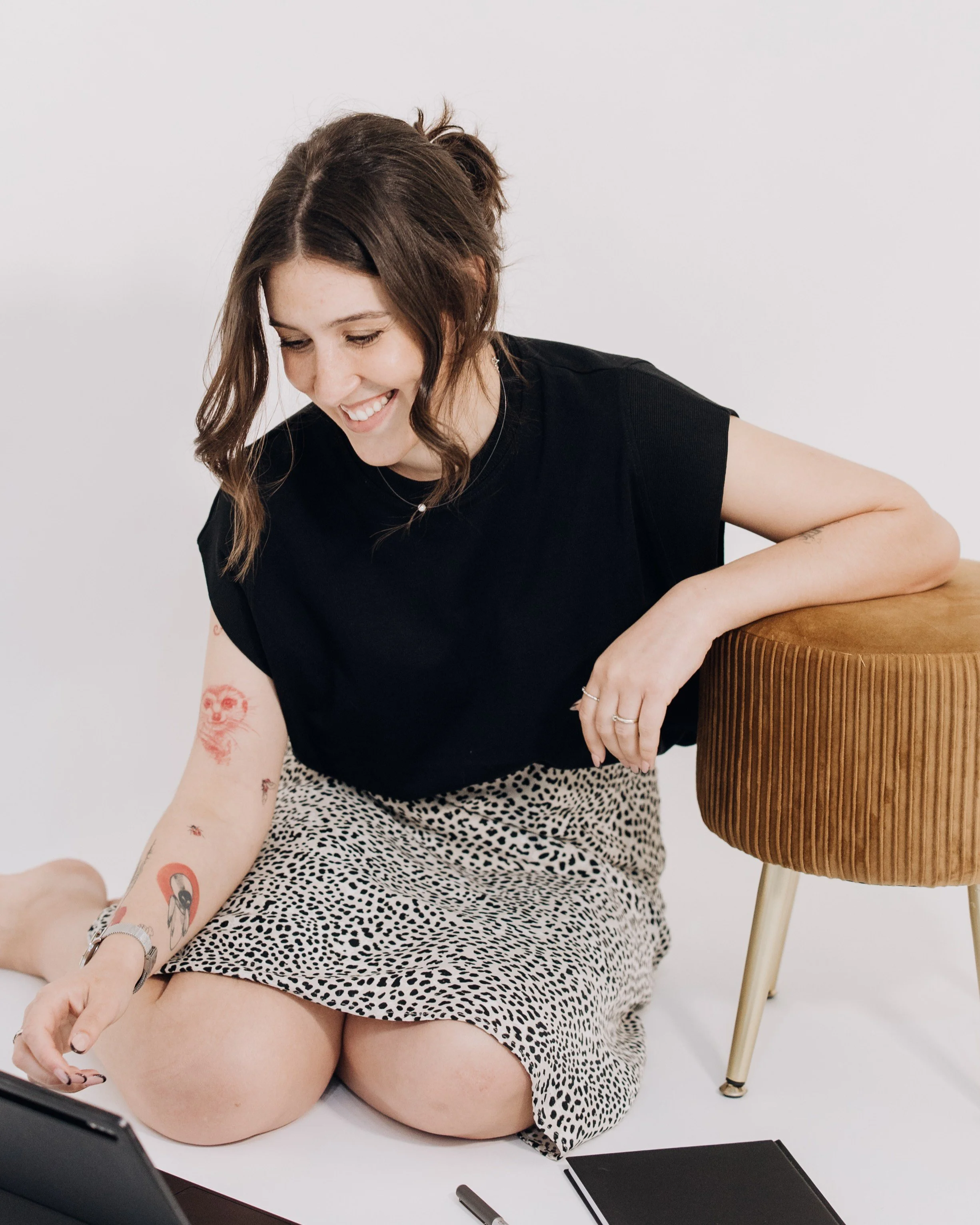 Pessoa sorridente com cabelo castanho, usando camiseta preta e saia de oncinha, sentada em uma cadeira de madeira, com tatuagens visíveis no braço e segurando um celular em uma mão. Ao seu lado há um caderno preto, uma caneta e atrás uma cadeira de madeira com pernas de metal.