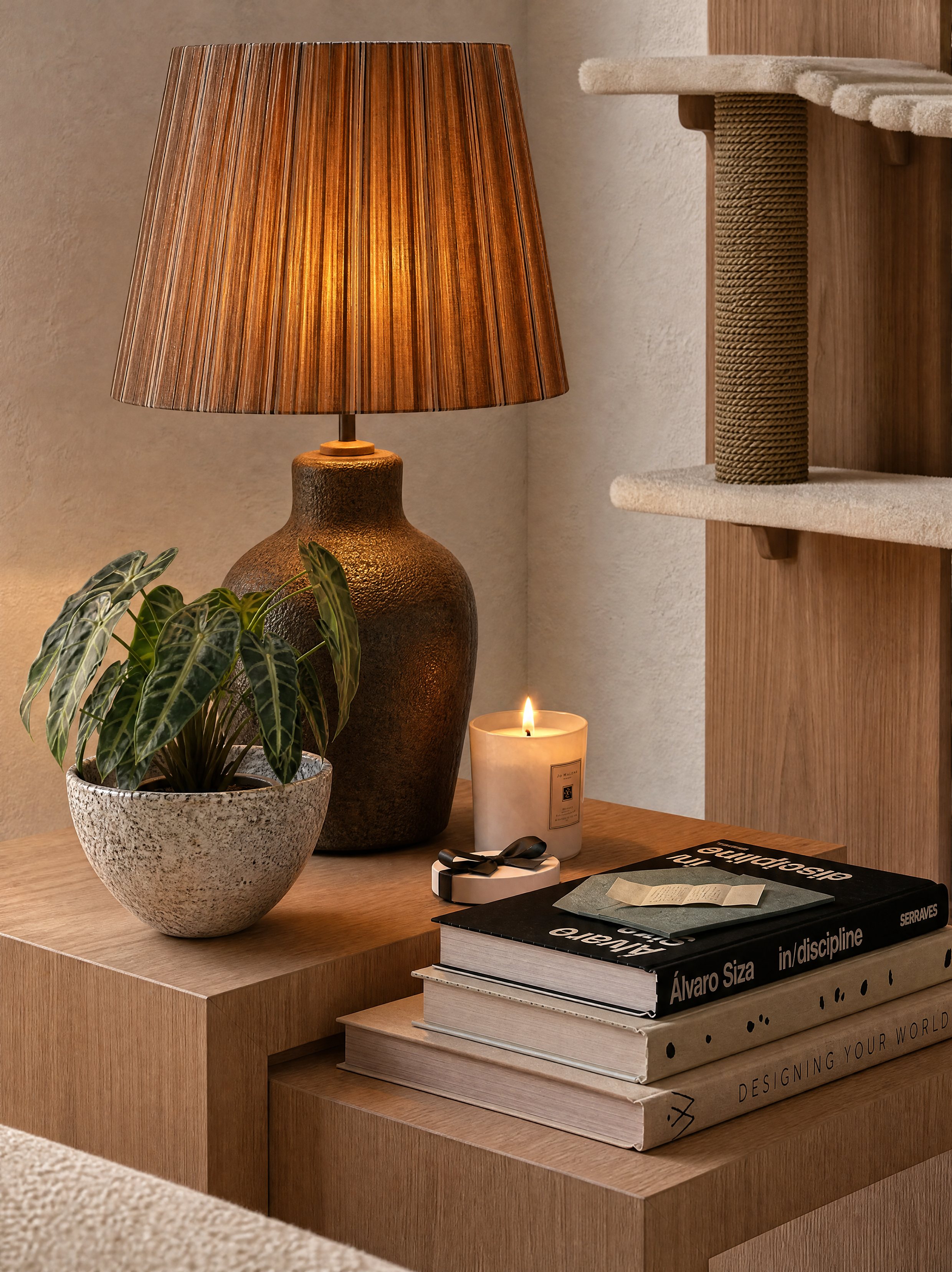 Mesa de madeira com vaso de planta com folhas verdes, abajur de madeira com cúpula listrada, vela acesa, e uma coleção de livros empilhados, incluindo títulos como 'in/discipline' e 'Designing Your World'.