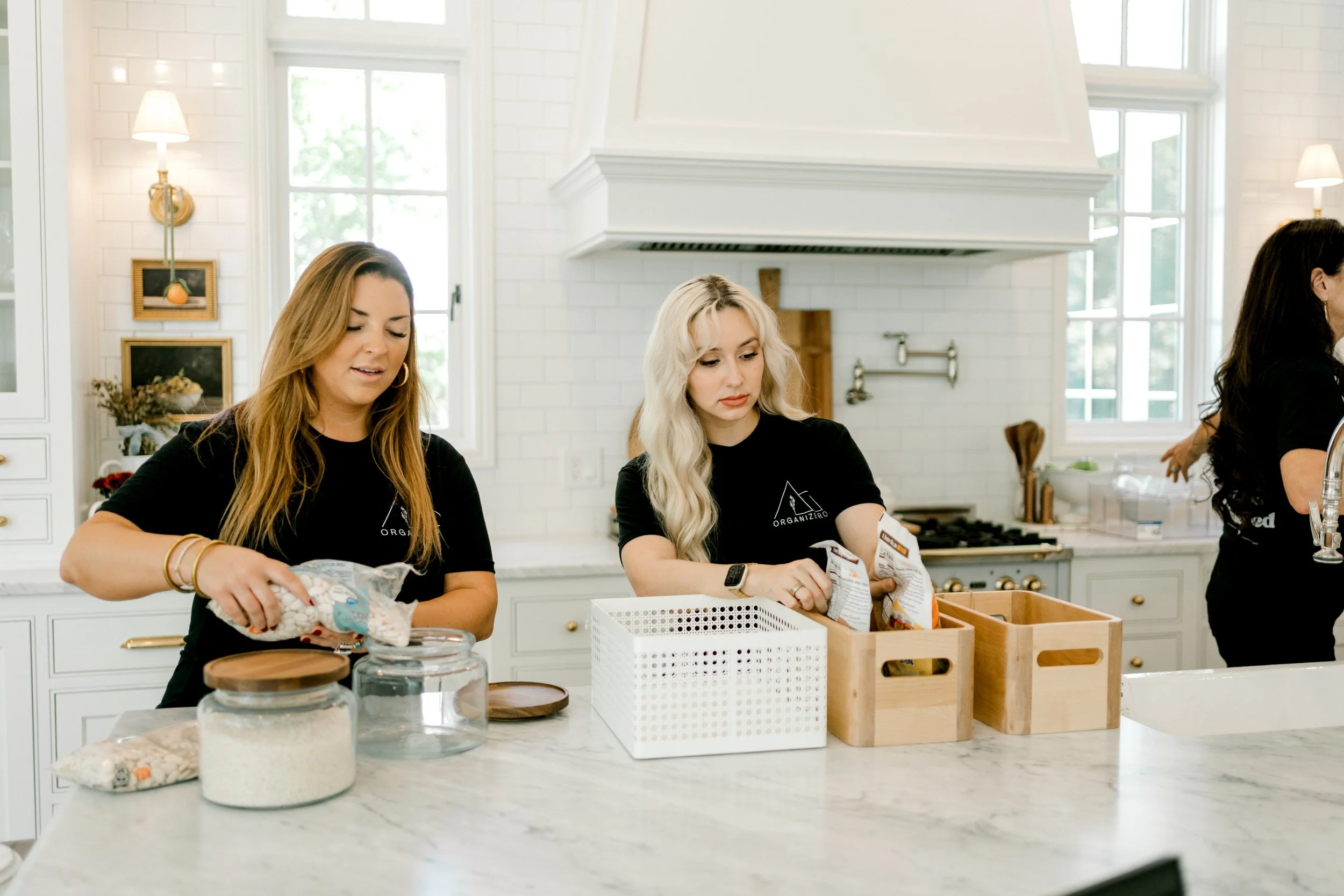Two OrganizePro professional organizers arranging pantry essentials in storage containers and jars in a bright kitchen.