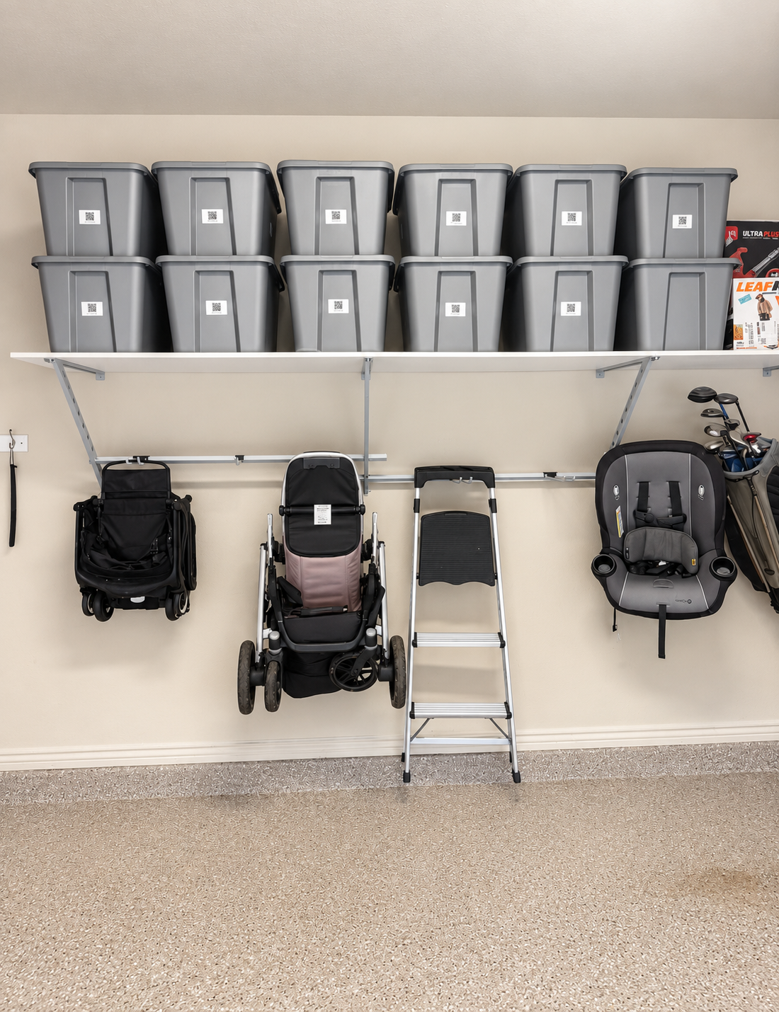 Organized garage with labeled gray storage bins on overhead shelving and strollers, a ladder, and baby equipment neatly stored on wall-mounted racks.