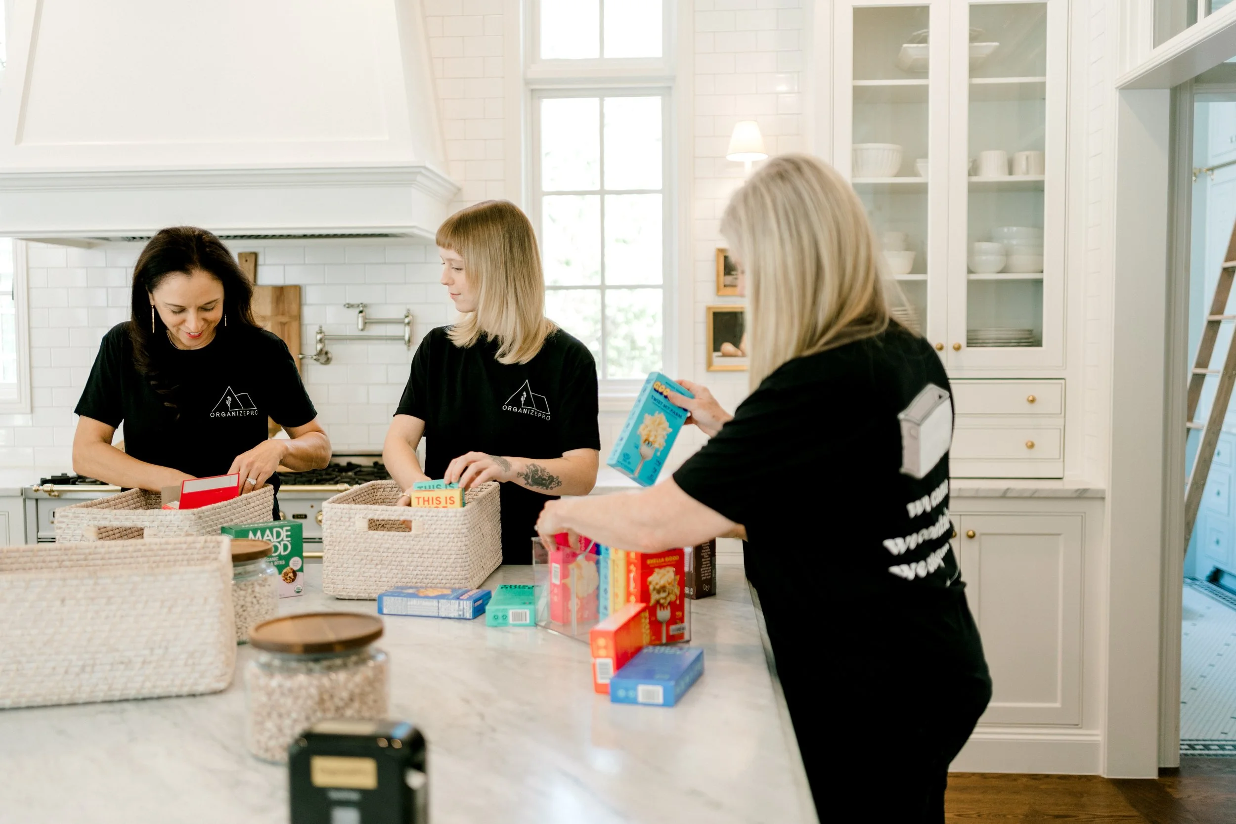 Three OrganizePro professional organizers working together in a bright kitchen, sorting pantry essentials and arranging food items into woven storage baskets.