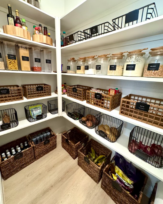 Walk-in pantry with labeled food storage jars, woven baskets, and black wire bins arranged on white shelves for easy access.