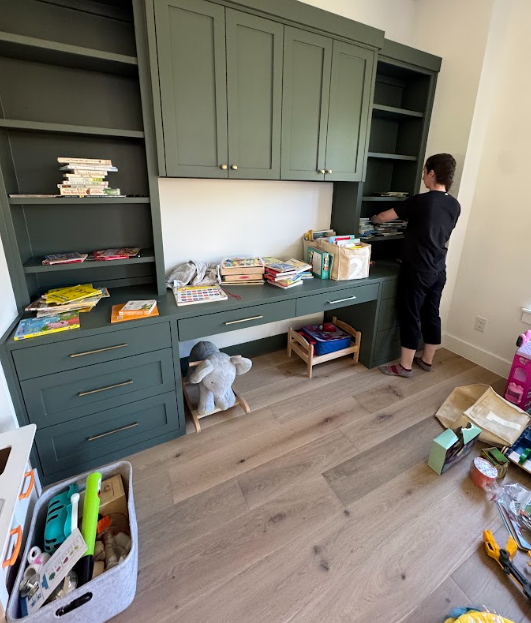 Professional organizer sorting children's books and toys on built-in green shelves while creating an organized playroom.