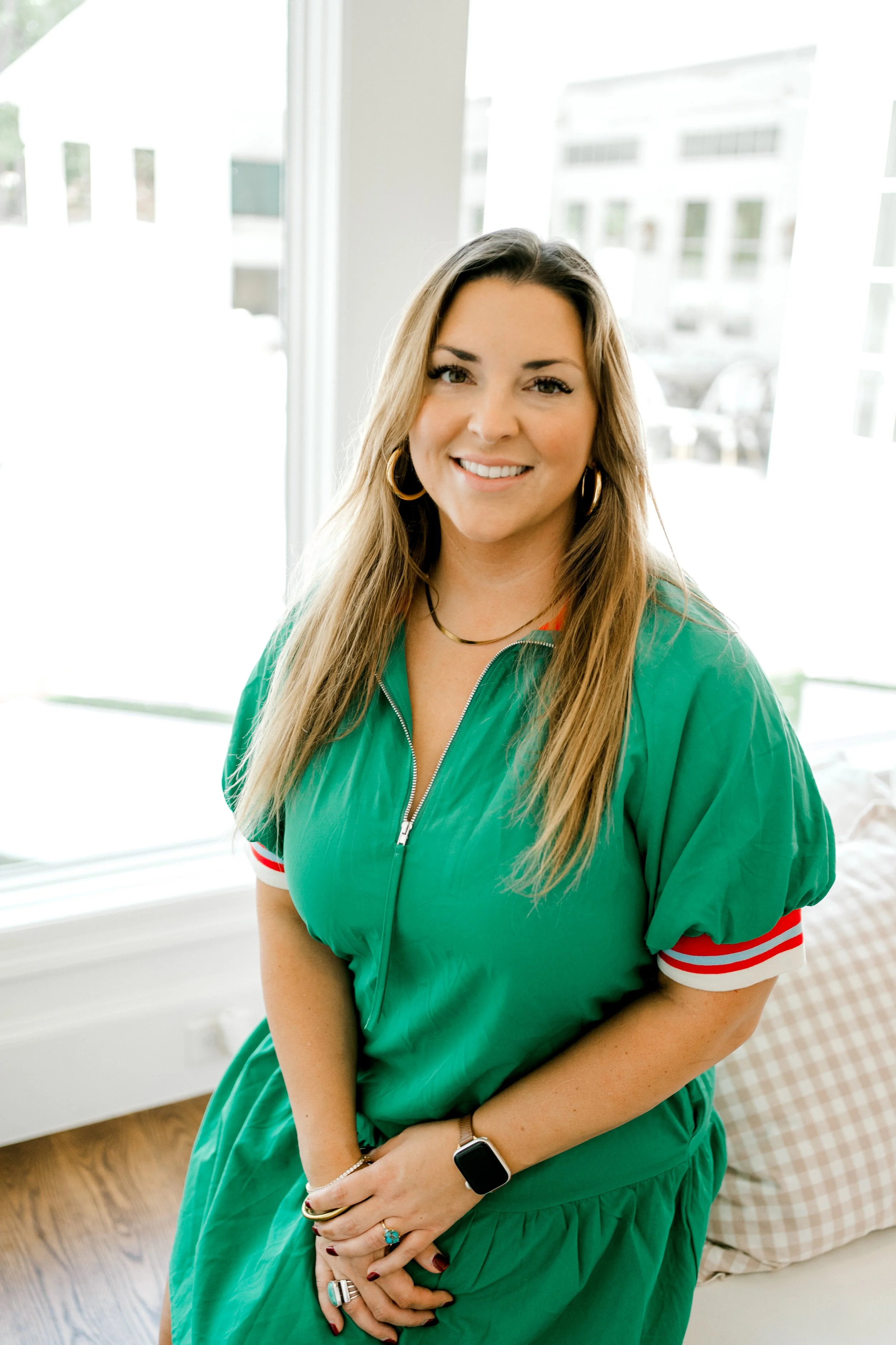Chandler, professional home organizer at OrganizePro, smiling in a green blouse while seated beside a bright window.