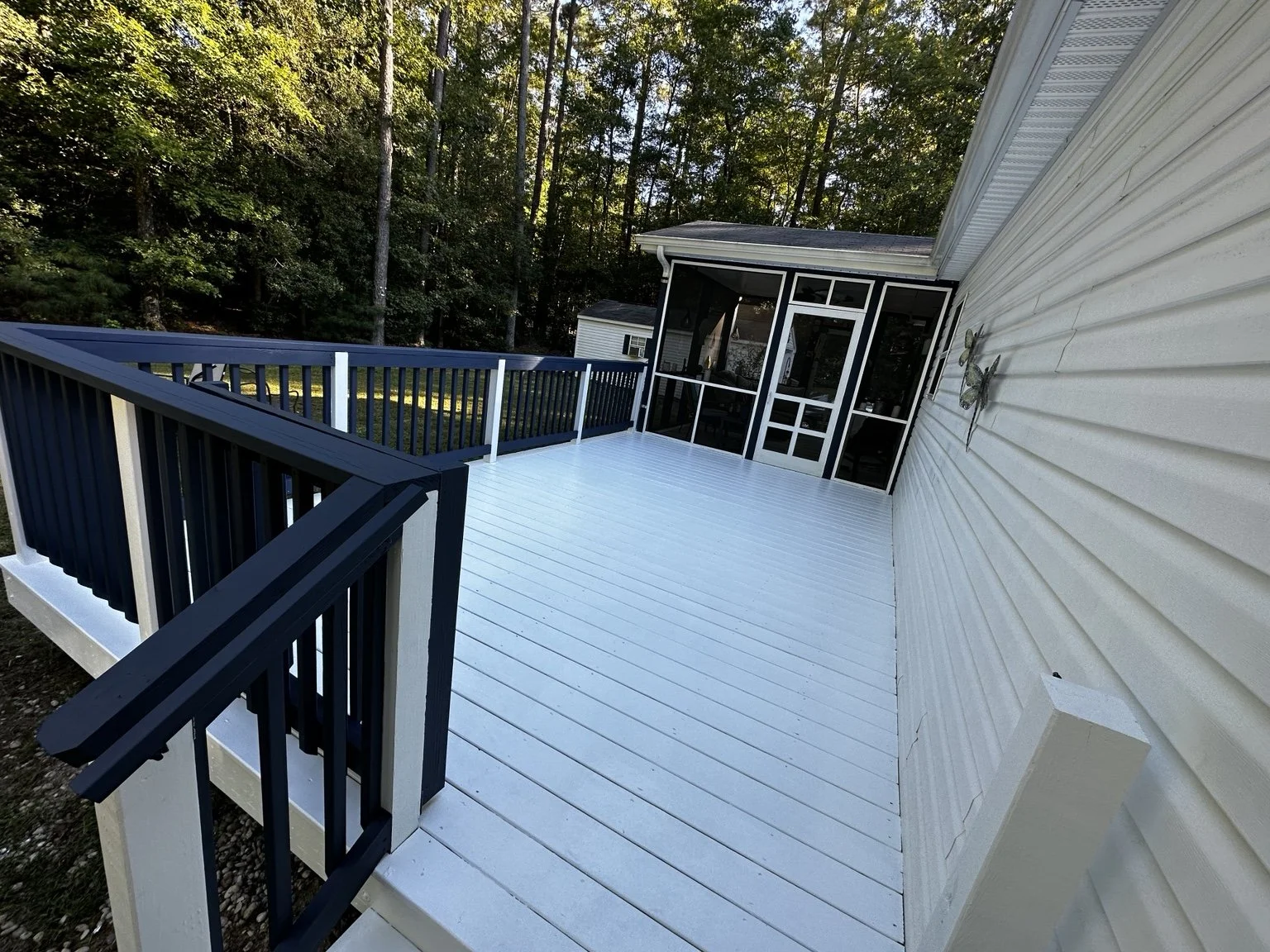 Empty outdoor balcony with white wooden floor, black and white railing, and a screened-in porch with a glass door, overlooking green trees.
