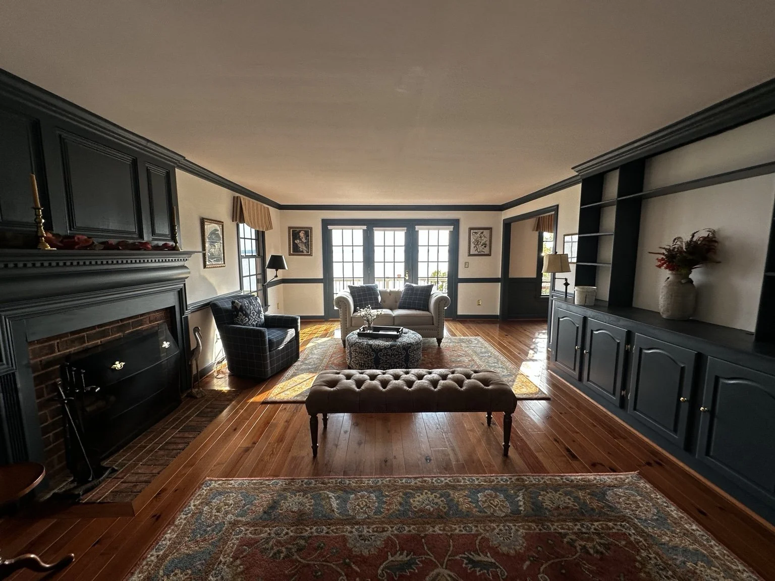 Living room with black built-in shelves, a white sofa, a black and white armchair, a tufted brown ottoman, a fireplace, wooden floors, and large windows.