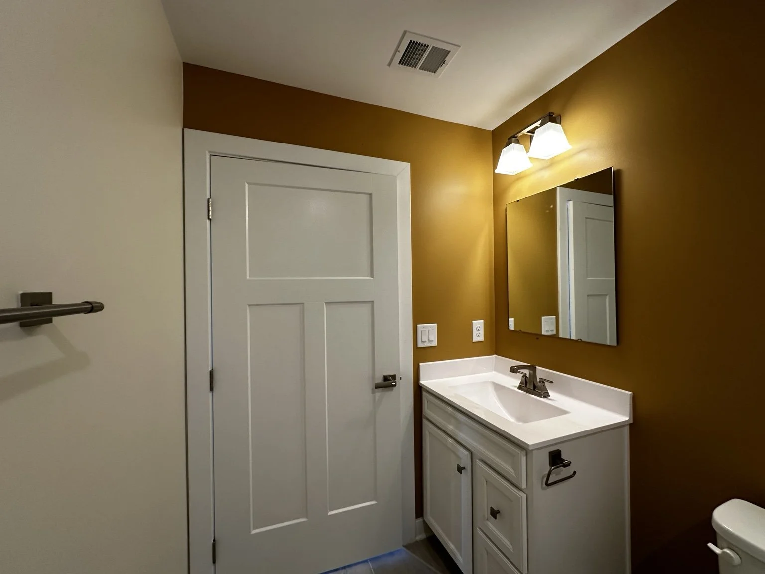 Bathroom with white door, wall-mounted towel bar, small white vanity with sink, mirror, and two lights above, mustard-colored wall, vent on ceiling, part of toilet visible.