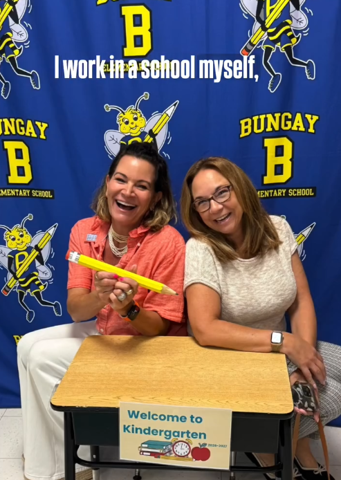 Two women sitting at a desk in front of a blue backdrop with the Bungay Elementary School logo, smiling and holding a giant pencil. There is a sign on the desk that says 'Welcome to Kindergarten'.