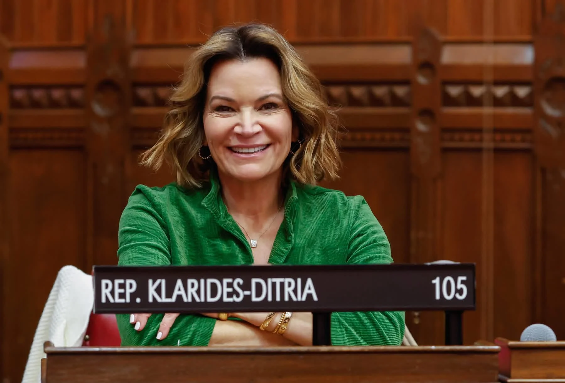 A woman with shoulder-length wavy hair smiling, wearing a green shirt and jewelry, sitting at a desk with a nameplate that reads "REP. KLARIDES-DITTRIA 105," in a formal setting with wooden paneling in the background.