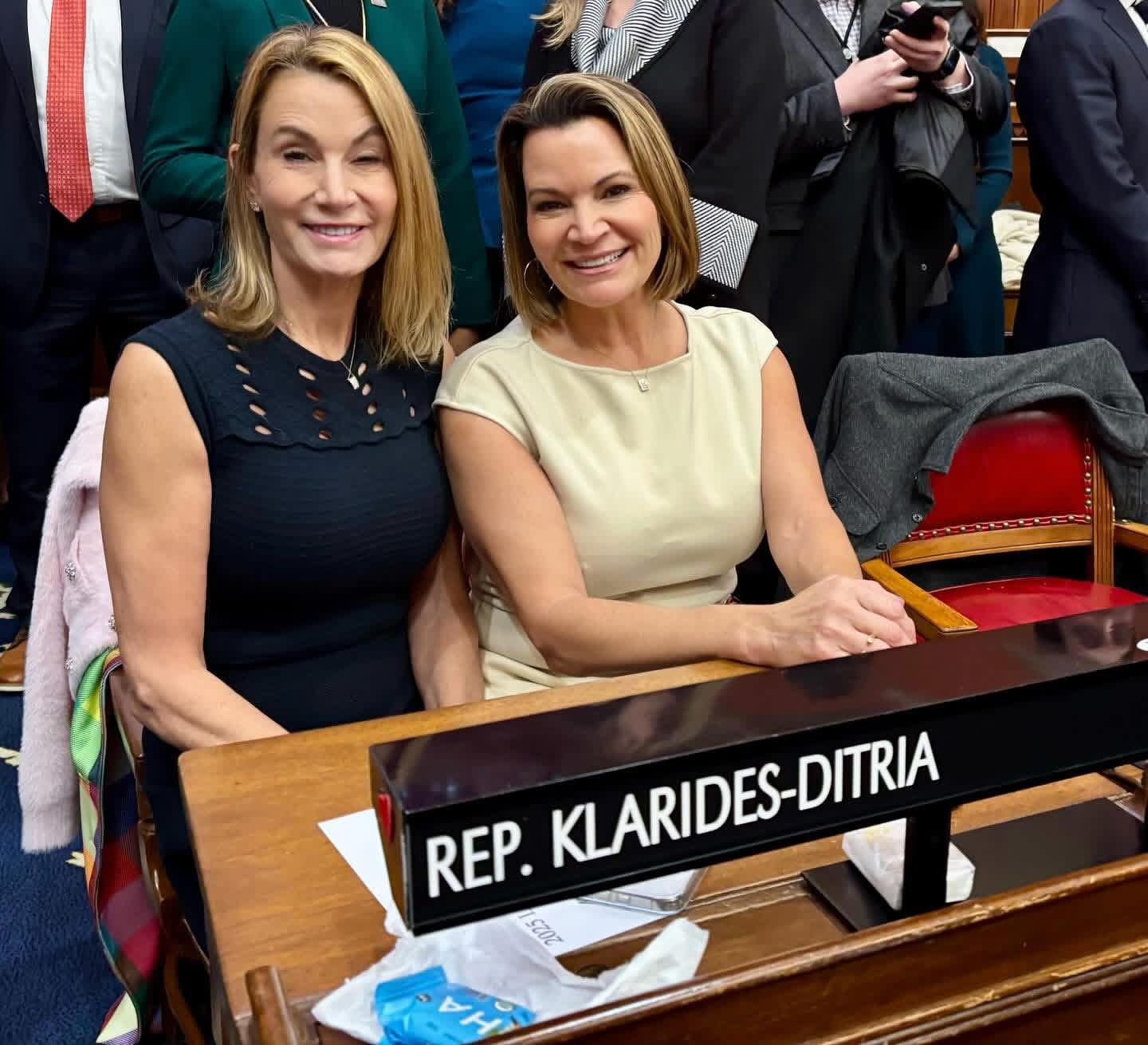 Two women sitting at a desk with a nameplate that reads 'Rep. Klarides-Ditria,' smiling at the camera, surrounded by people in a formal setting.