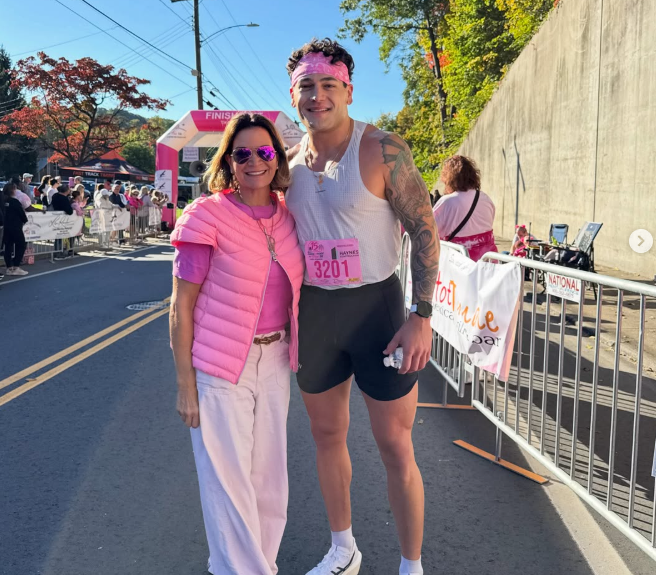 A woman and a man smiling at a finish line at a race event on a sunny day, with spectators and a pink finish arch in the background.