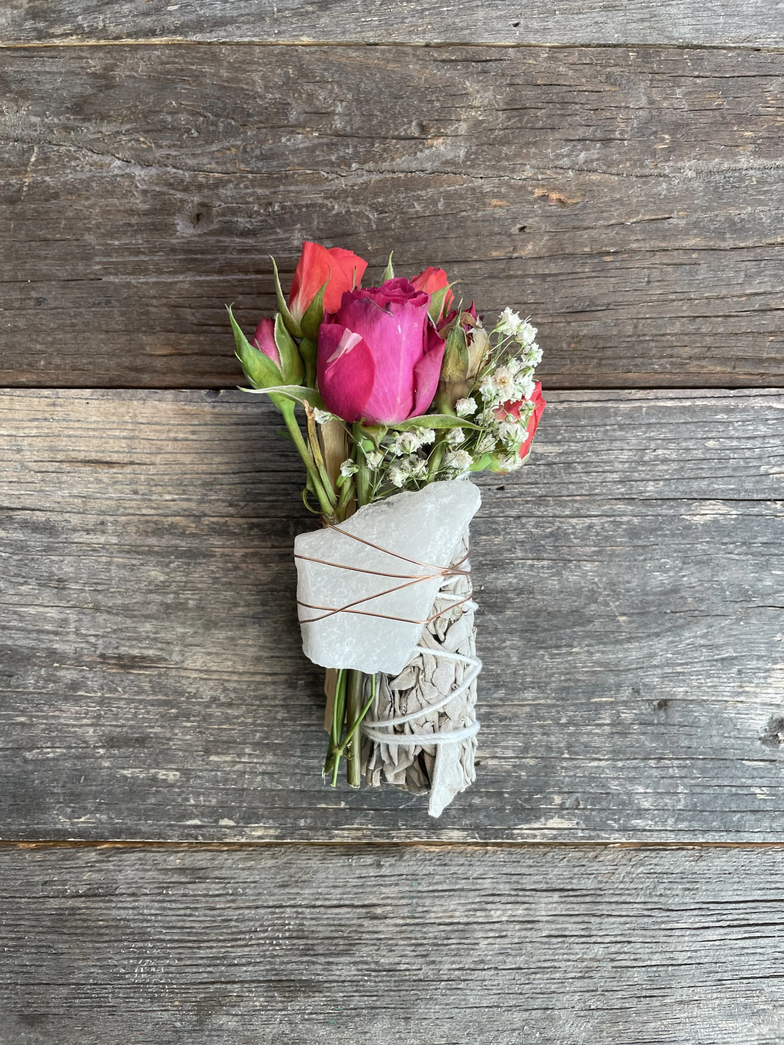 A small bouquet of pink and red roses with white baby's breath flowers, wrapped in white fabric and tied with copper wire, placed on a rustic wooden surface.