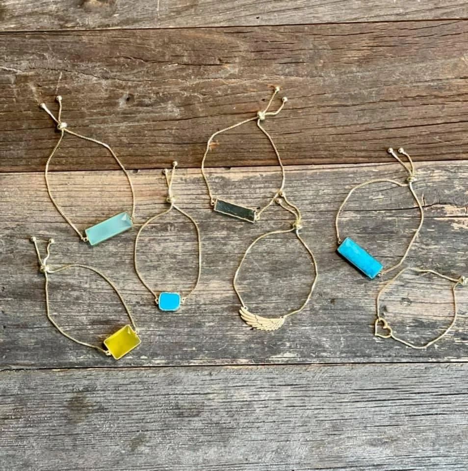 Six gold-colored bracelets with colorful rectangular and heart-shaped charms are laid out on a wooden surface.