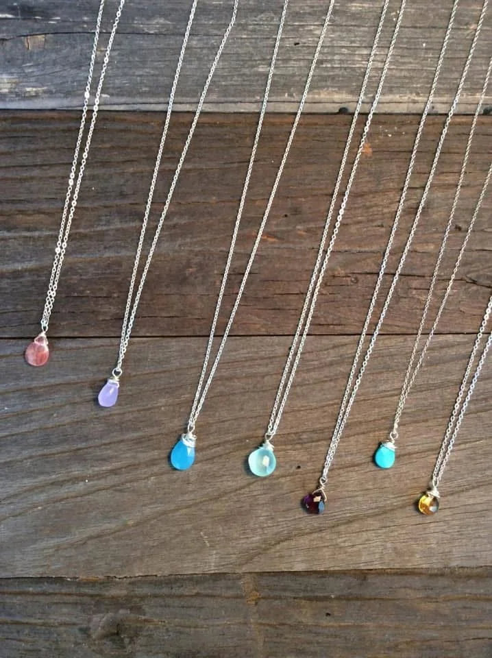 Six necklaces with colorful teardrop-shaped pendants arranged in a row on a wooden surface.