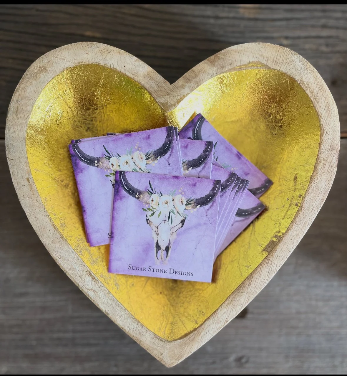 Heart-shaped wooden box with gold interior holding several business cards featuring a bull skull with flowers, labeled 'Sugar Stone Designs'.