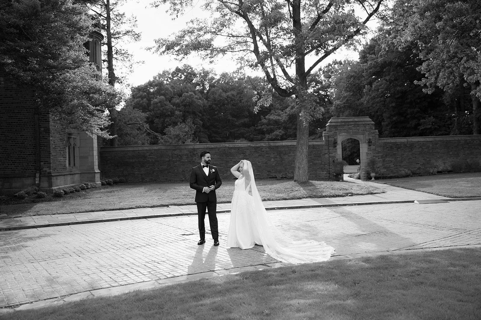 Black-and-white wedding portrait of a bride and groom standing outside a brick estate wall and garden entrance