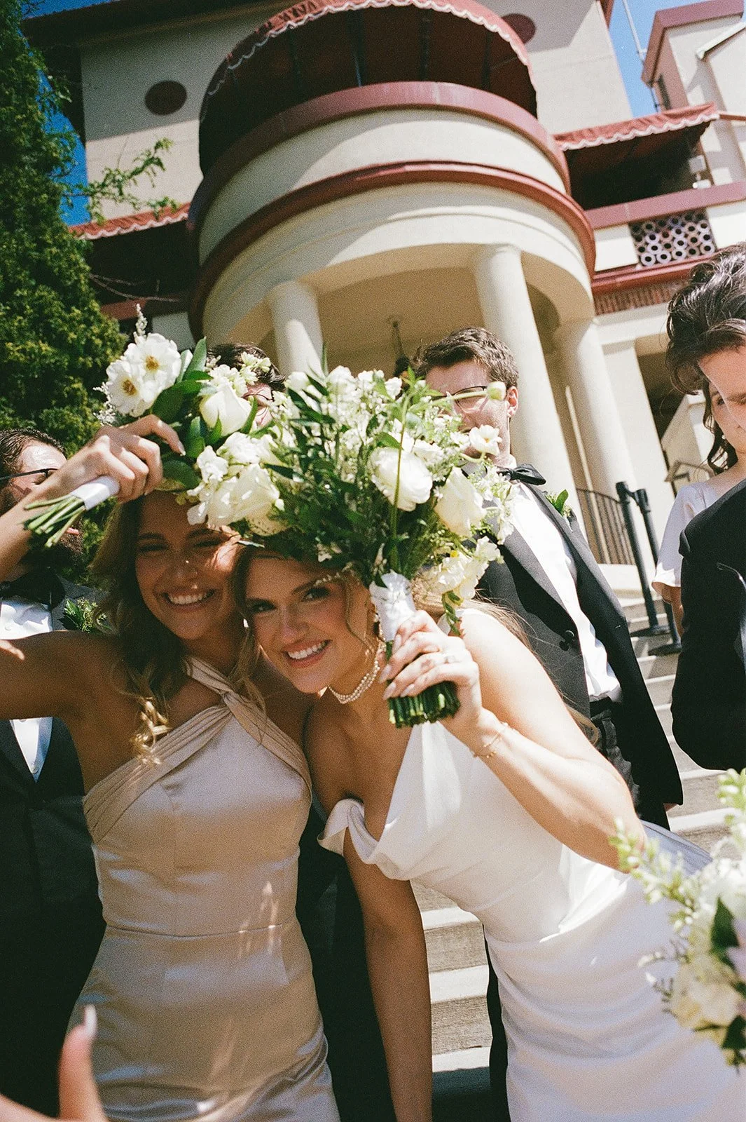 Smiling bride holding a white floral bouquet with her bridesmaid and wedding guests outside a building