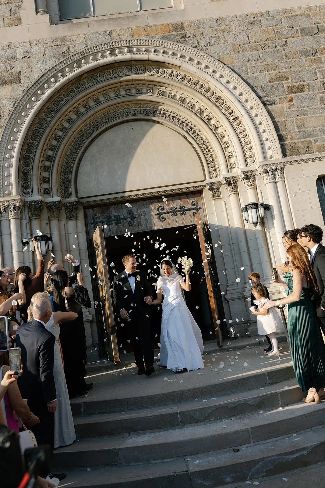 Newlywed couple leaving an ornate stone church as guests toss flower petals