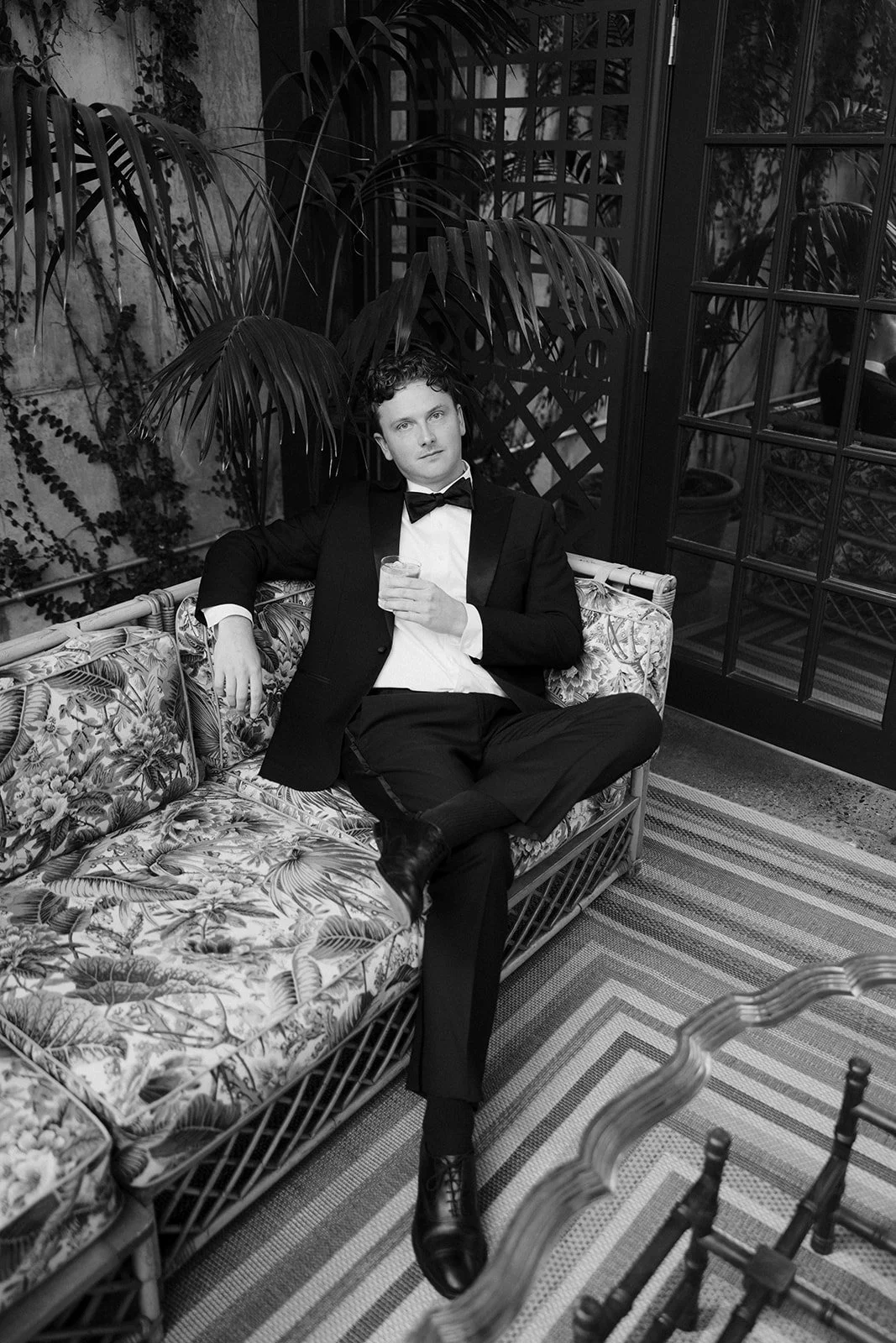 Black-and-white portrait of a man in a tuxedo sitting on a tropical floral-patterned wicker sofa, holding a drink.