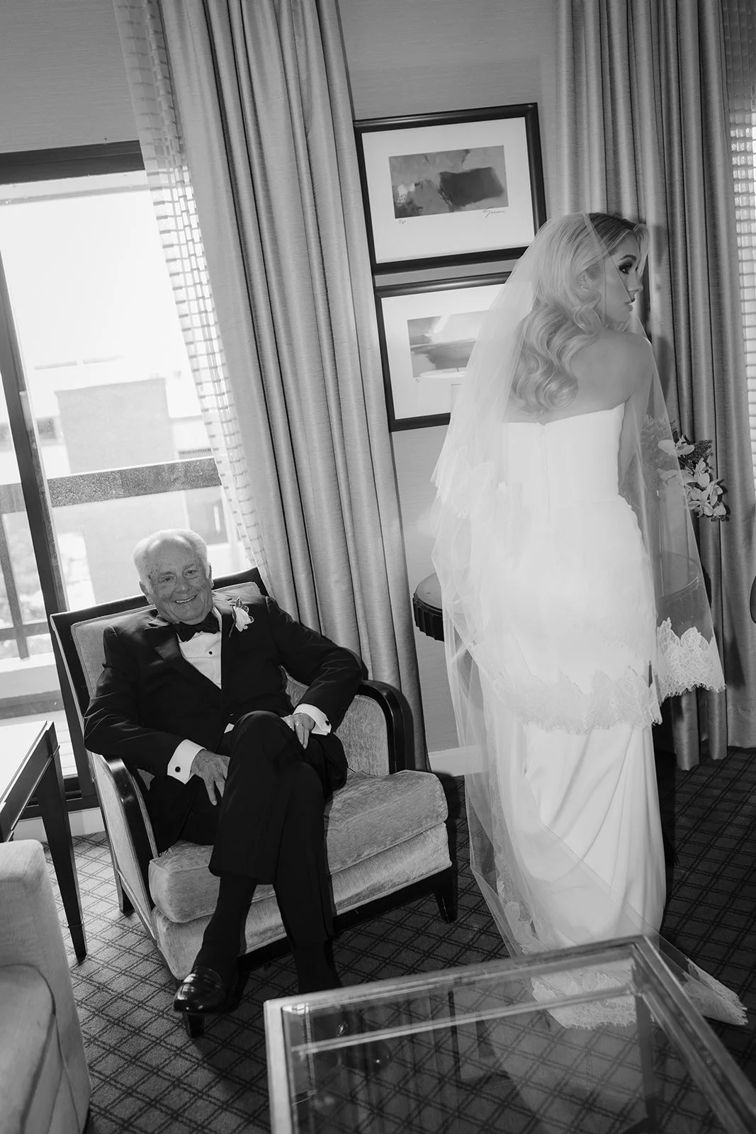 Black-and-white wedding portrait of a bride in a veil standing beside an older man in a tuxedo seated in an armchair.