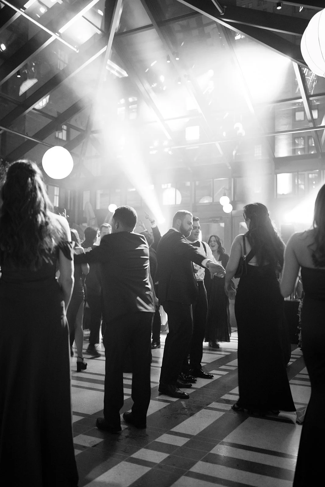 Black-and-white photo of formally dressed guests dancing at an elegant indoor reception with dramatic lighting.