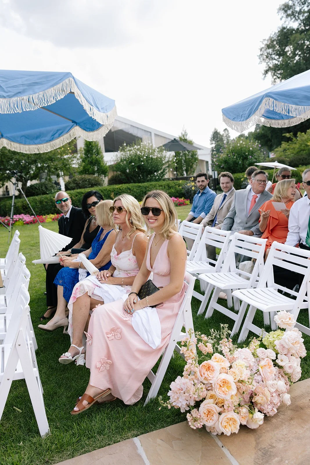 Wedding guests seated on white chairs beneath blue-fringed umbrellas in a garden ceremony setting, with pastel flowers in the foreground