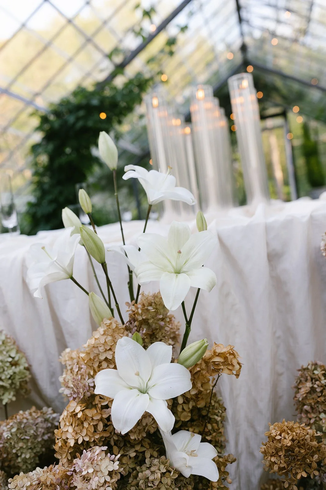 White lilies and dried hydrangeas arranged in a sunlit greenhouse with hanging lights and white drapery