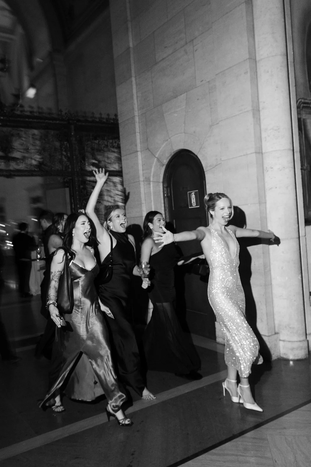 Group of women laughing and posing in elegant evening gowns outside a grand building.