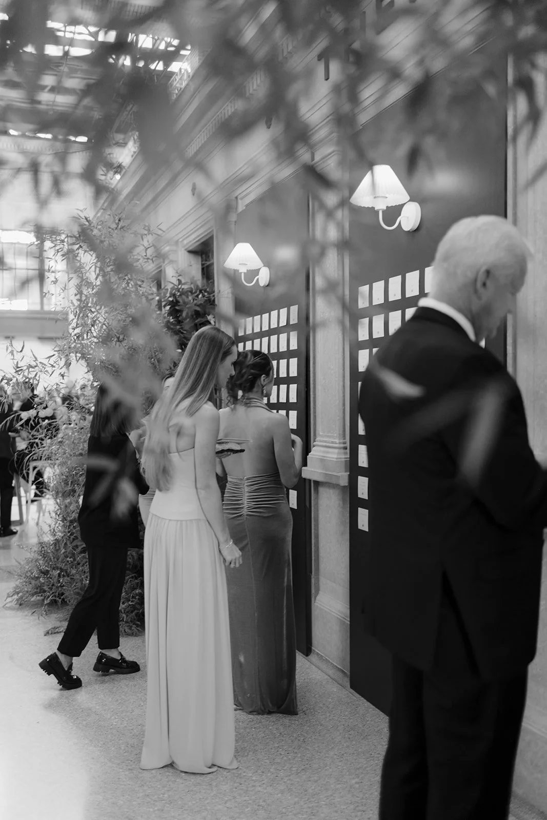 Guests in formal attire view seating cards at an elegant indoor event venue.