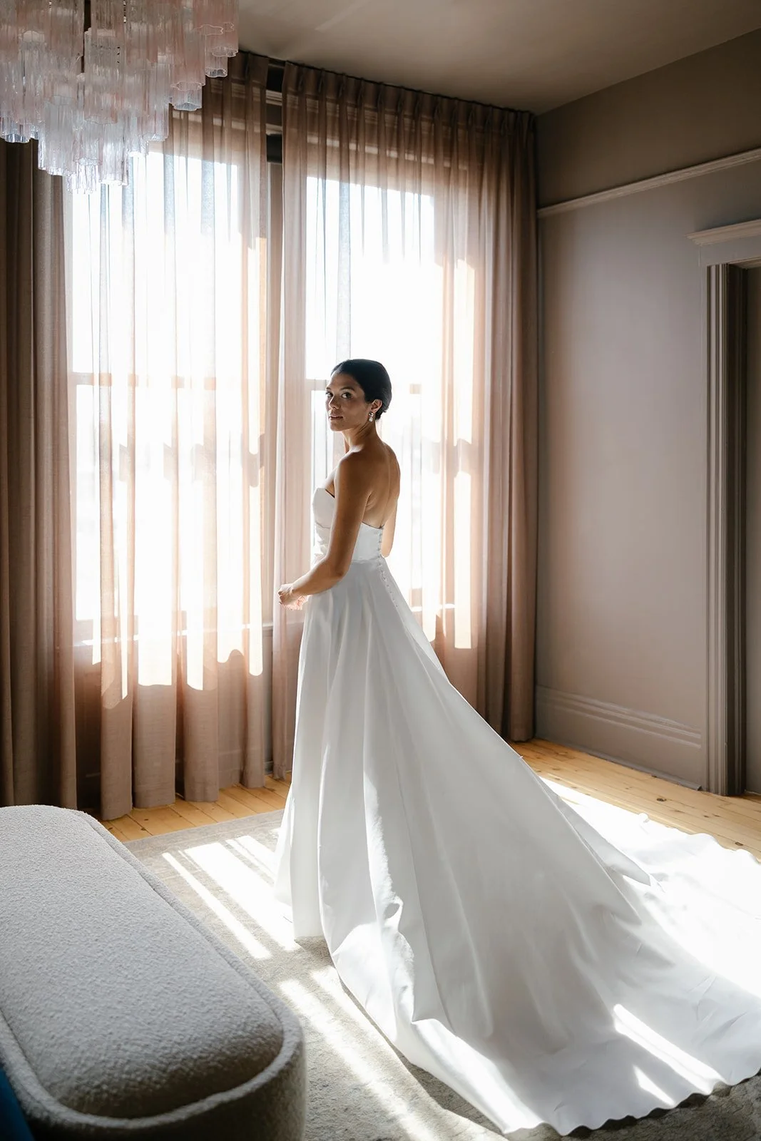 Bride in a strapless white wedding gown standing in a sunlit room with floor-to-ceiling curtains