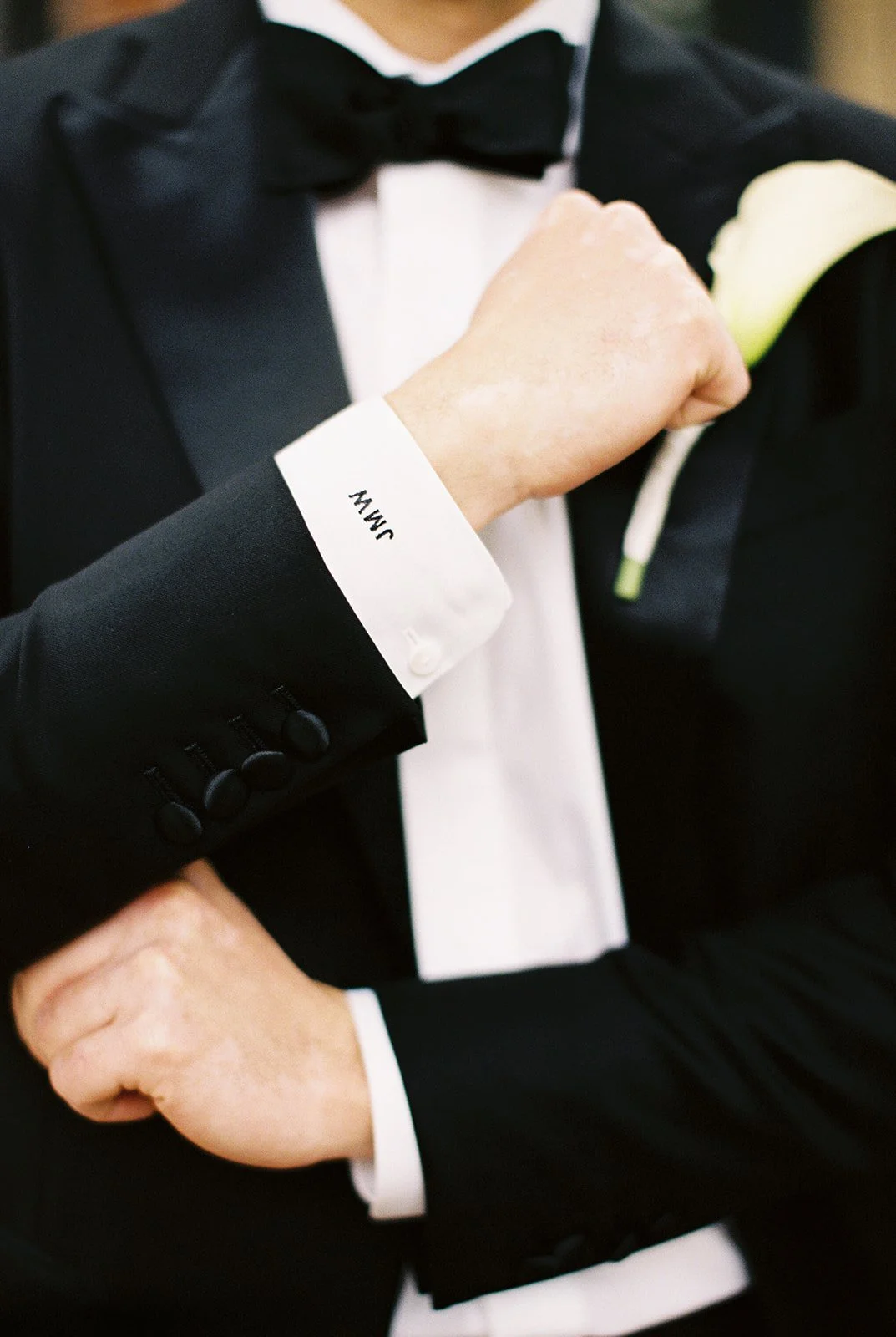 Close-up of a groom in a black tuxedo adjusting his cufflink beside a white boutonniere.