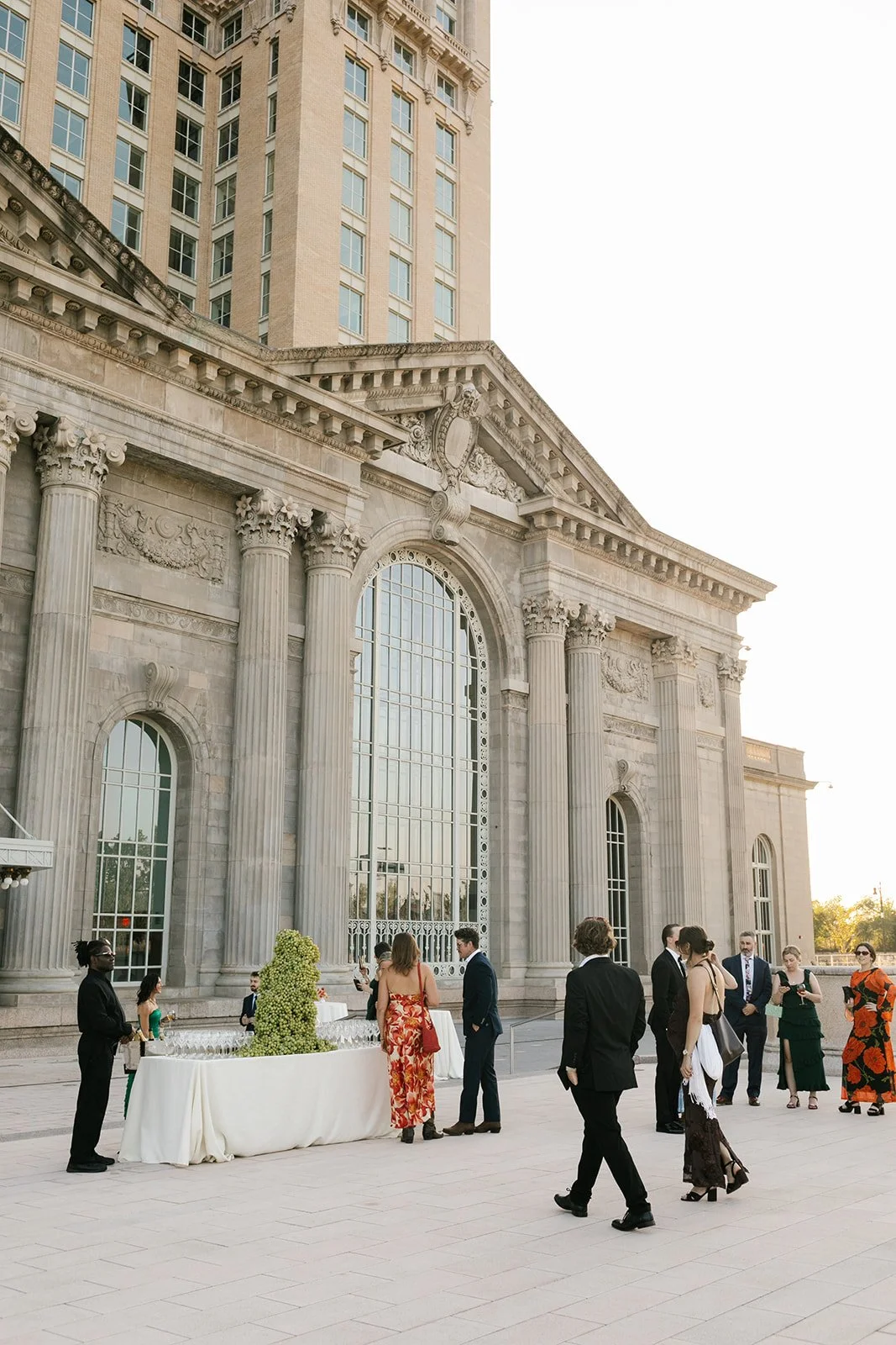 Guests gather at an outdoor reception beside an ornate classical building, with a white table displaying champagne glasses and a green grape tower.