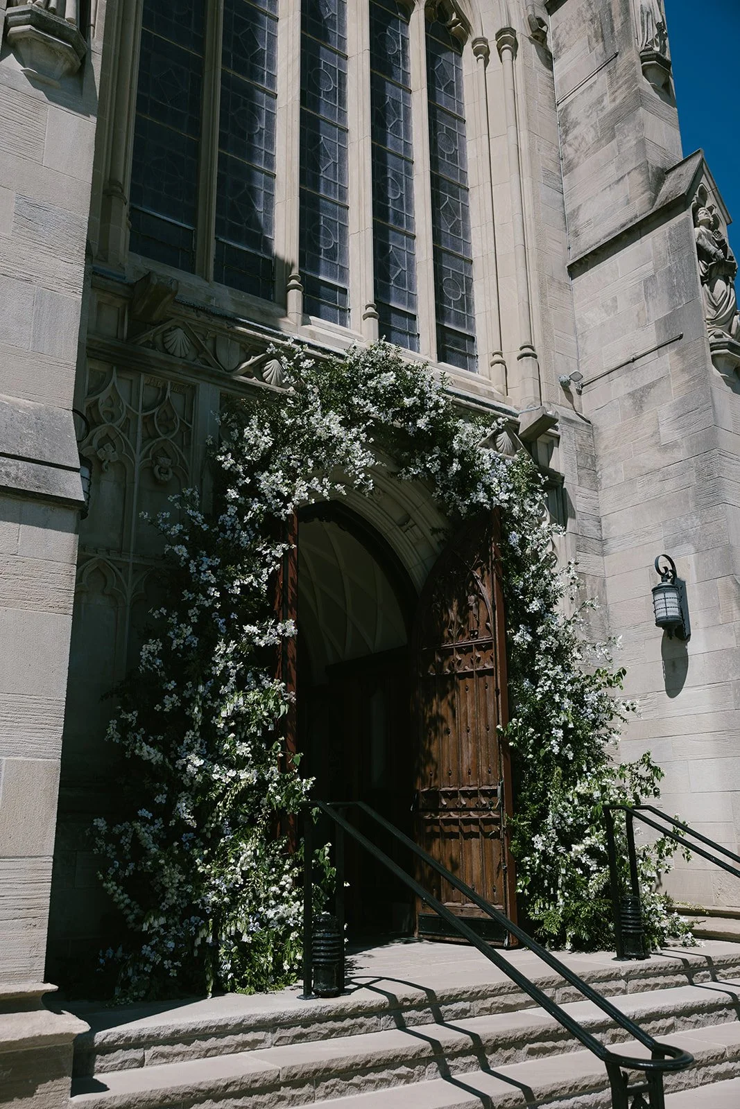 Gothic stone church entrance with open wooden doors, stained-glass windows, carved statues, and white flowering vines