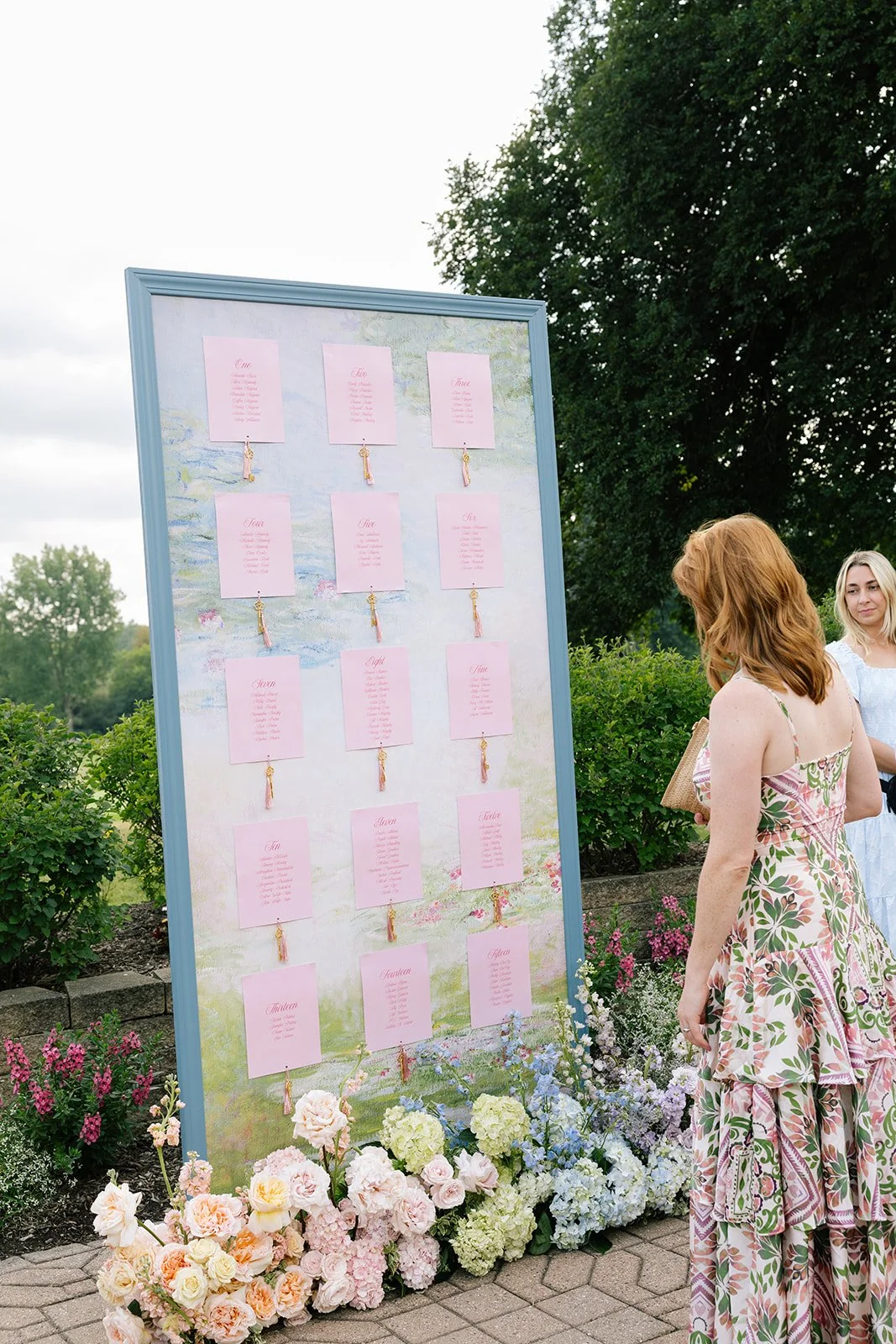 Guests view a pastel floral wedding seating chart displayed outdoors among colorful garden flowers.