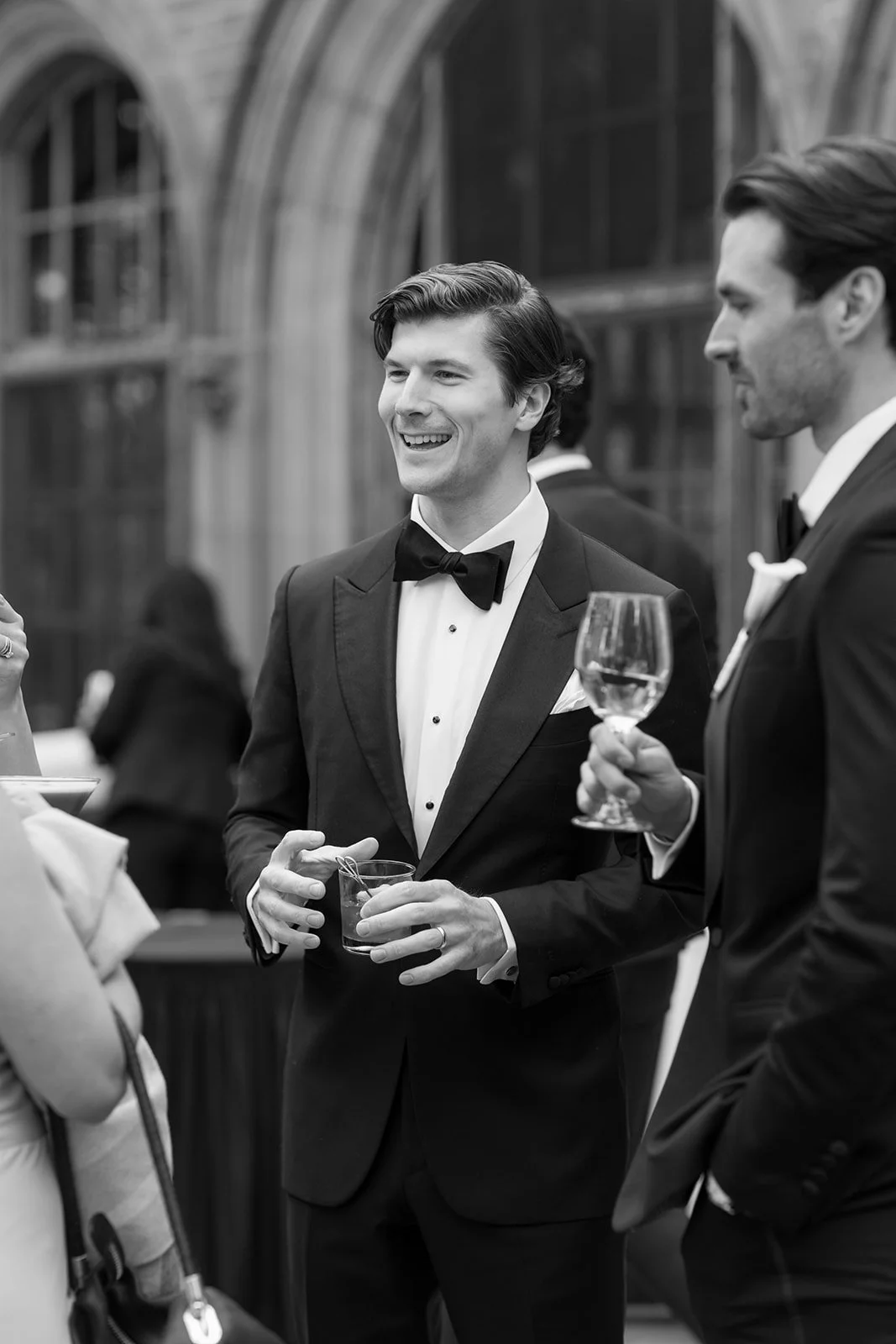 Black-and-white photo of smiling guests in formal tuxedos holding drinks at an elegant reception.