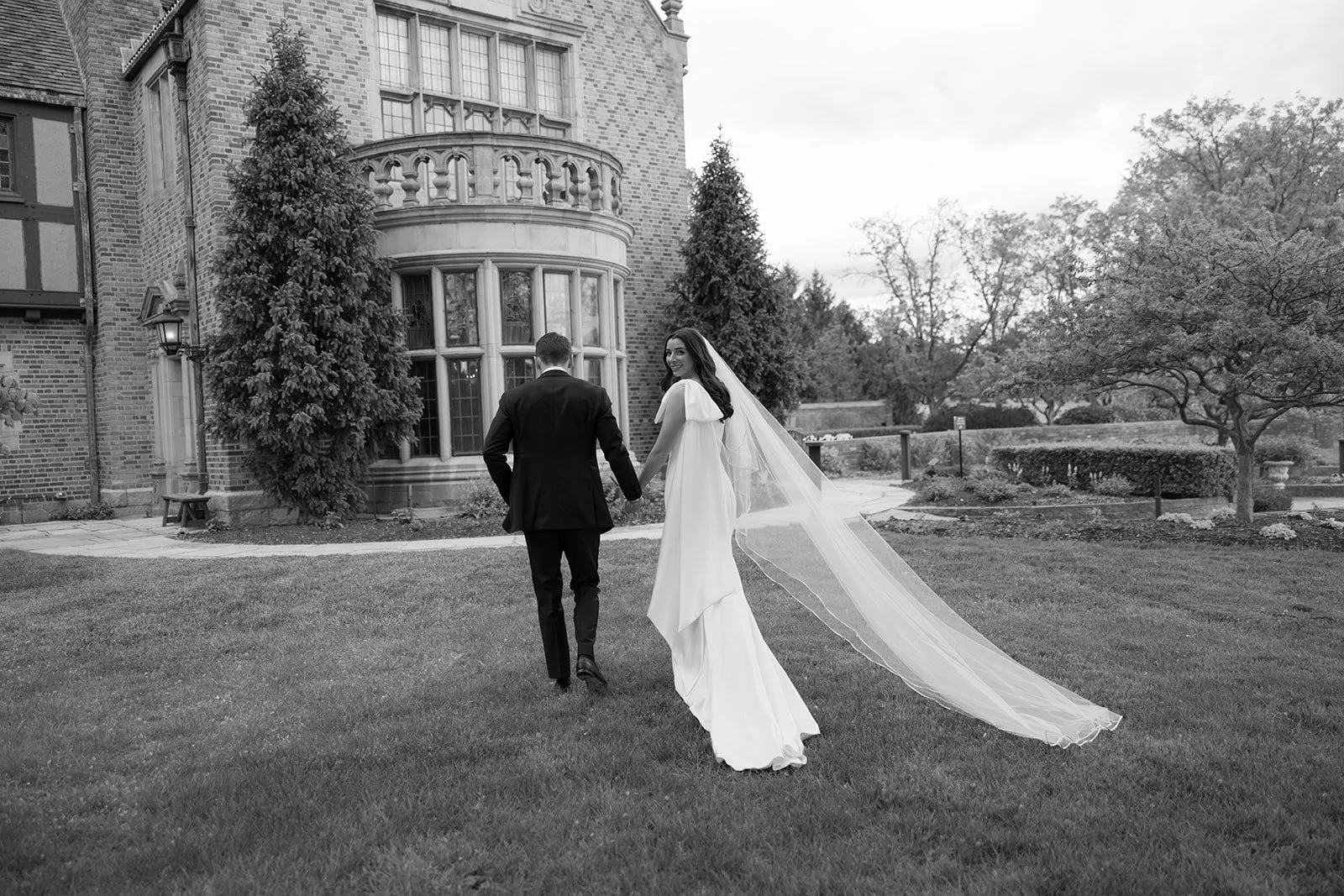 Bride and groom walking hand in hand across a lawn outside a historic brick manor house, with the bride’s long veil trailing behind
