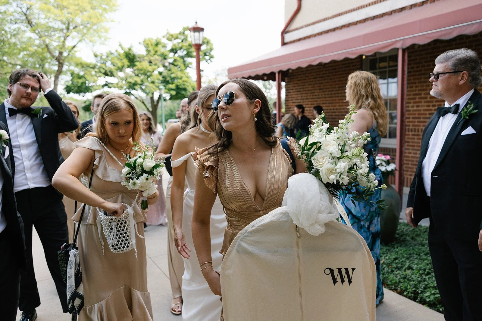 Wedding guests in formal attire gather outside a brick venue, with bouquets and a monogrammed chair.
