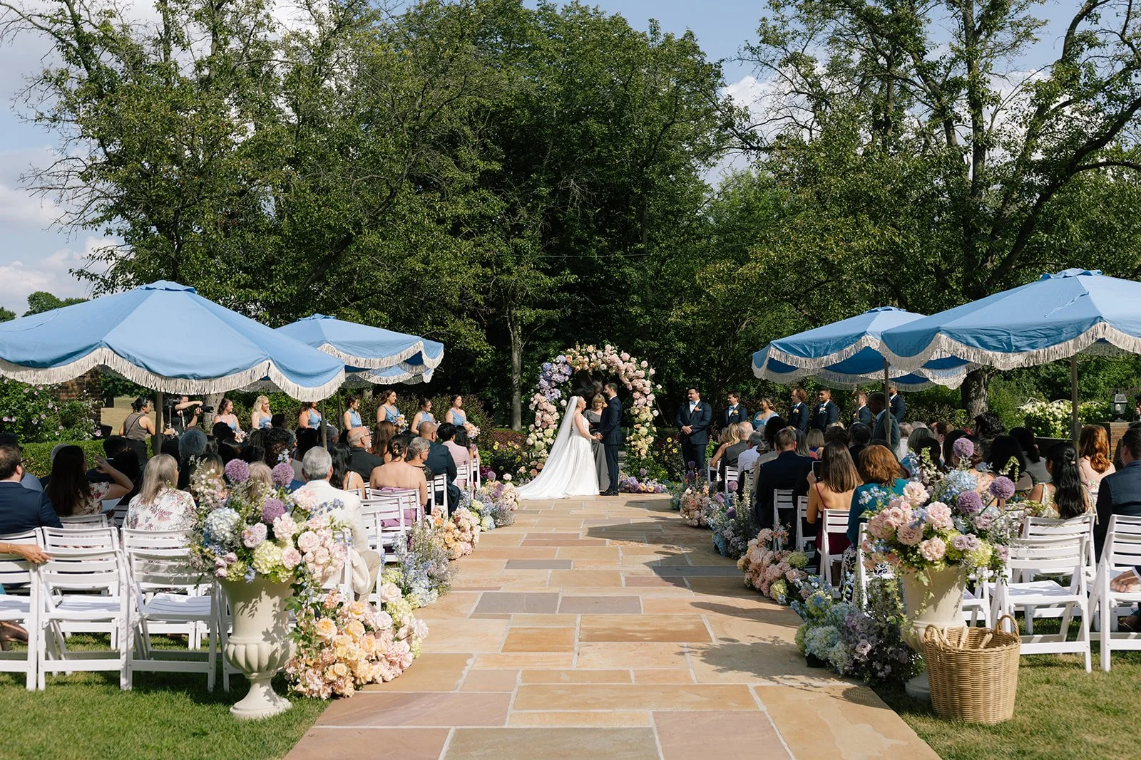 Outdoor garden wedding ceremony with bride and groom beneath a floral arch, guests seated under blue umbrellas, and pastel flower arrangements
