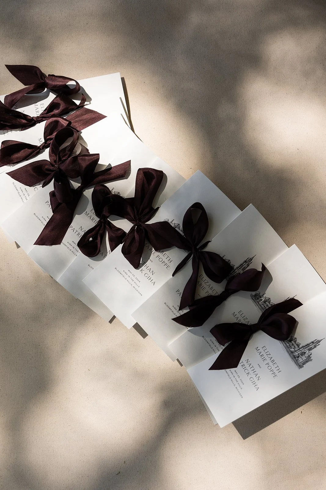 White wedding programs with dark burgundy satin bows arranged on a linen surface in sunlight