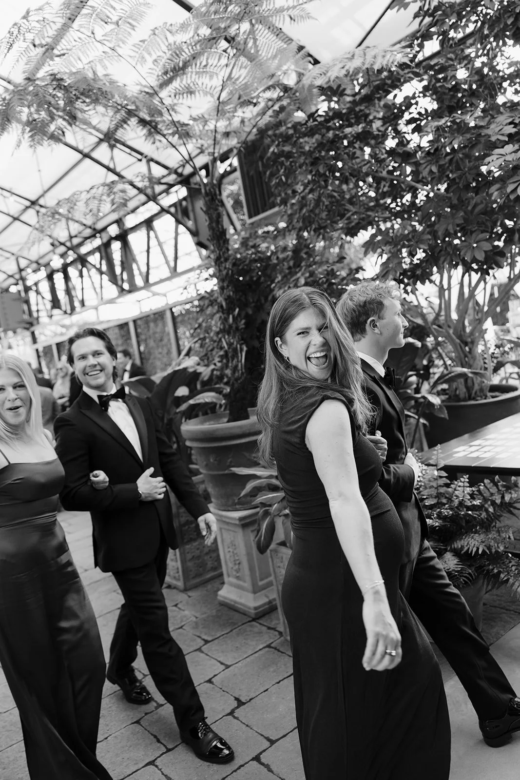 Guests in formal attire laughing and walking through a lush indoor conservatory at an event