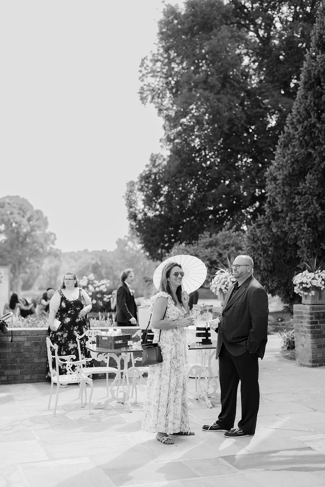 Guests mingle at an outdoor garden reception, with a woman holding a parasol and a man in a suit near decorated tables.