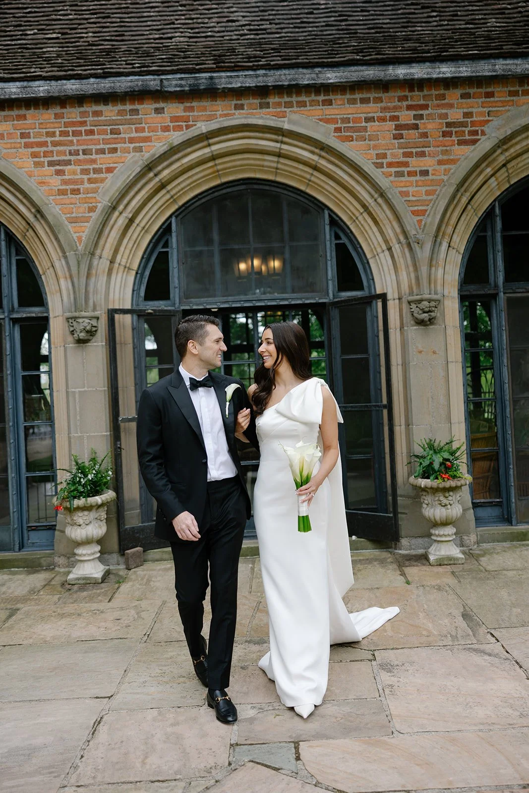 Newlywed couple posing outside a historic brick building, with the groom in a black tuxedo and the bride in a white wedding dress holding a bouquet.