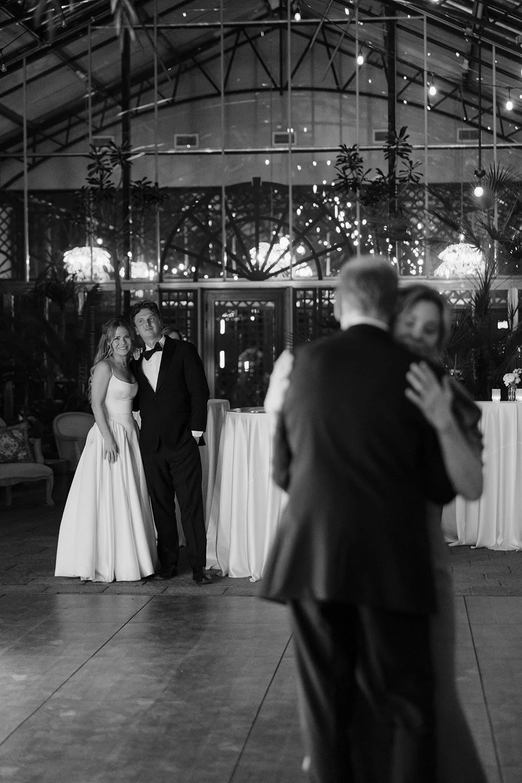 Black-and-white wedding reception photo with a bride and groom posing while another couple dances in the foreground.