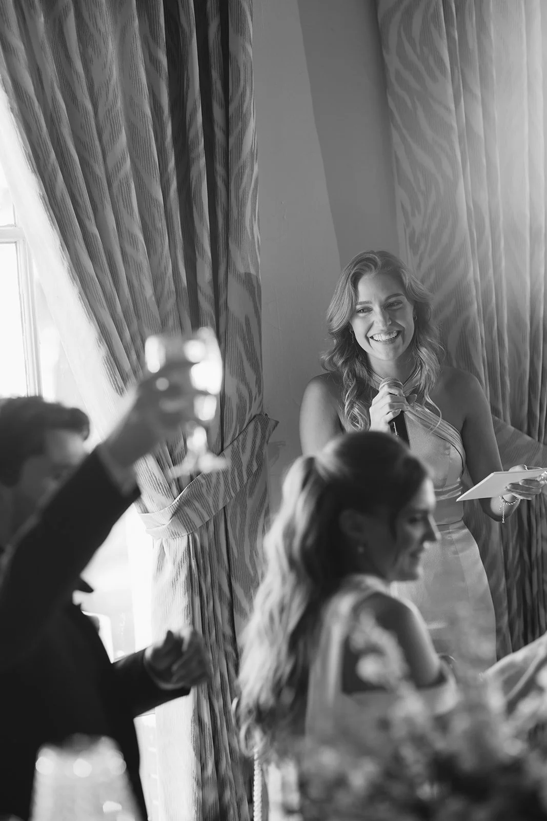 Smiling woman speaking into a microphone at an indoor event while guests take photos.