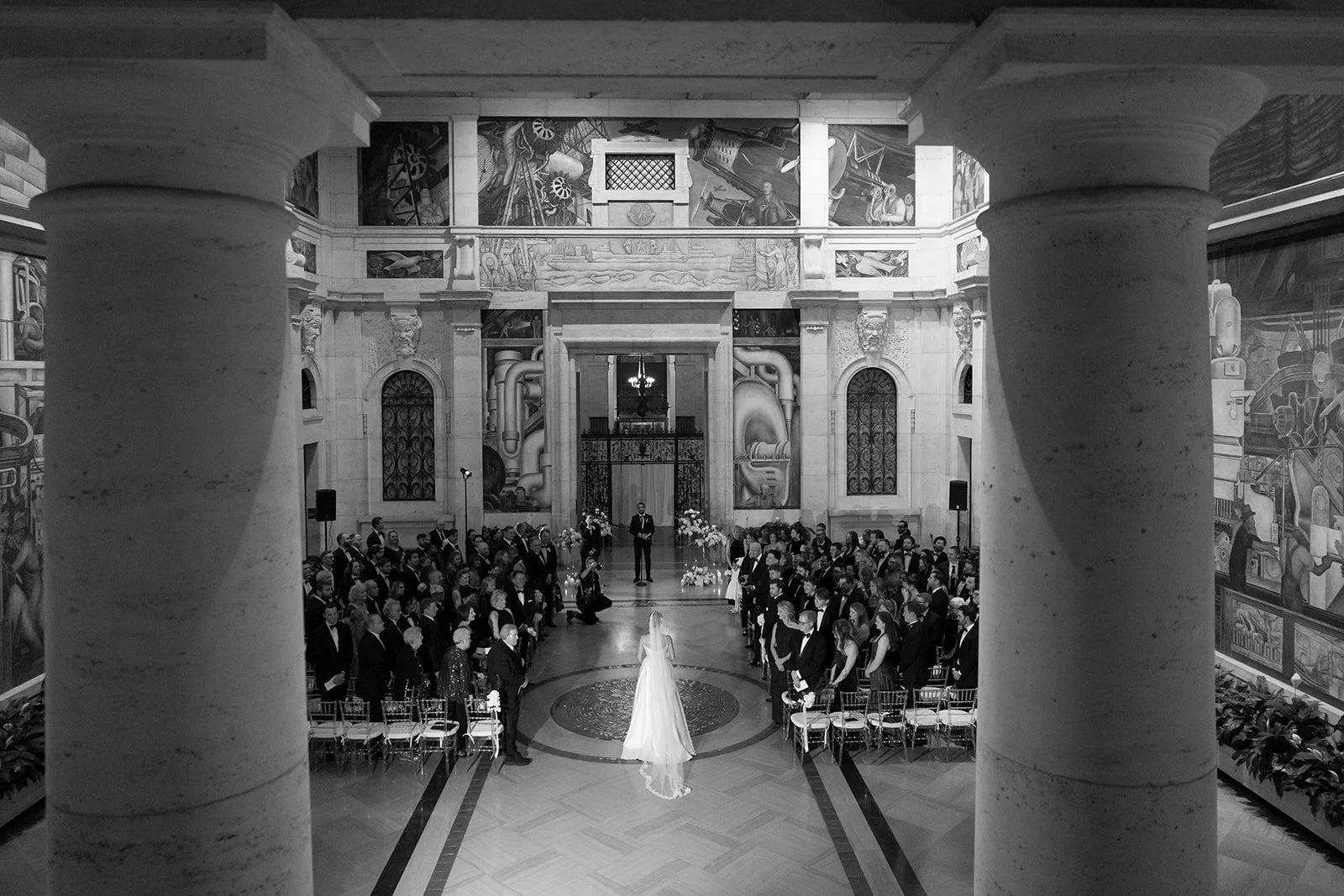 Black-and-white wedding ceremony in an ornate hall with murals, columns, bride, groom, and seated guests