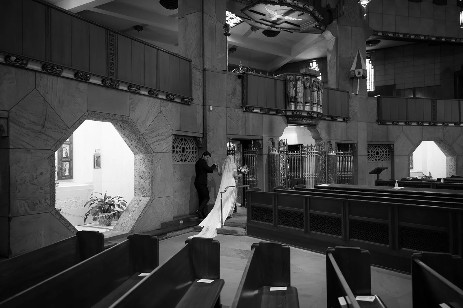 Black-and-white photo of a bride and groom inside an ornate church sanctuary with wooden pews and decorative marble architecture