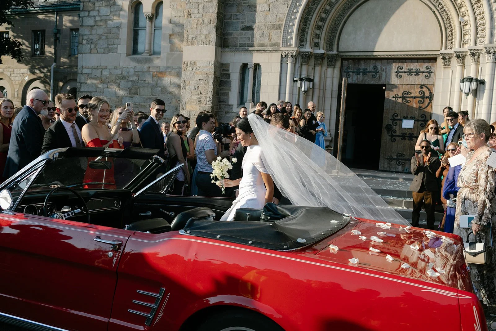 Bride in a white wedding dress and veil near a red vintage convertible outside a stone church, surrounded by wedding guests.