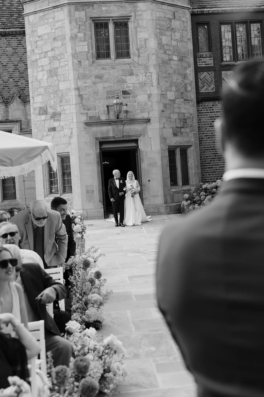 Bride and groom emerge from a historic stone building during an outdoor wedding ceremony with seated guests and floral aisle decorations.