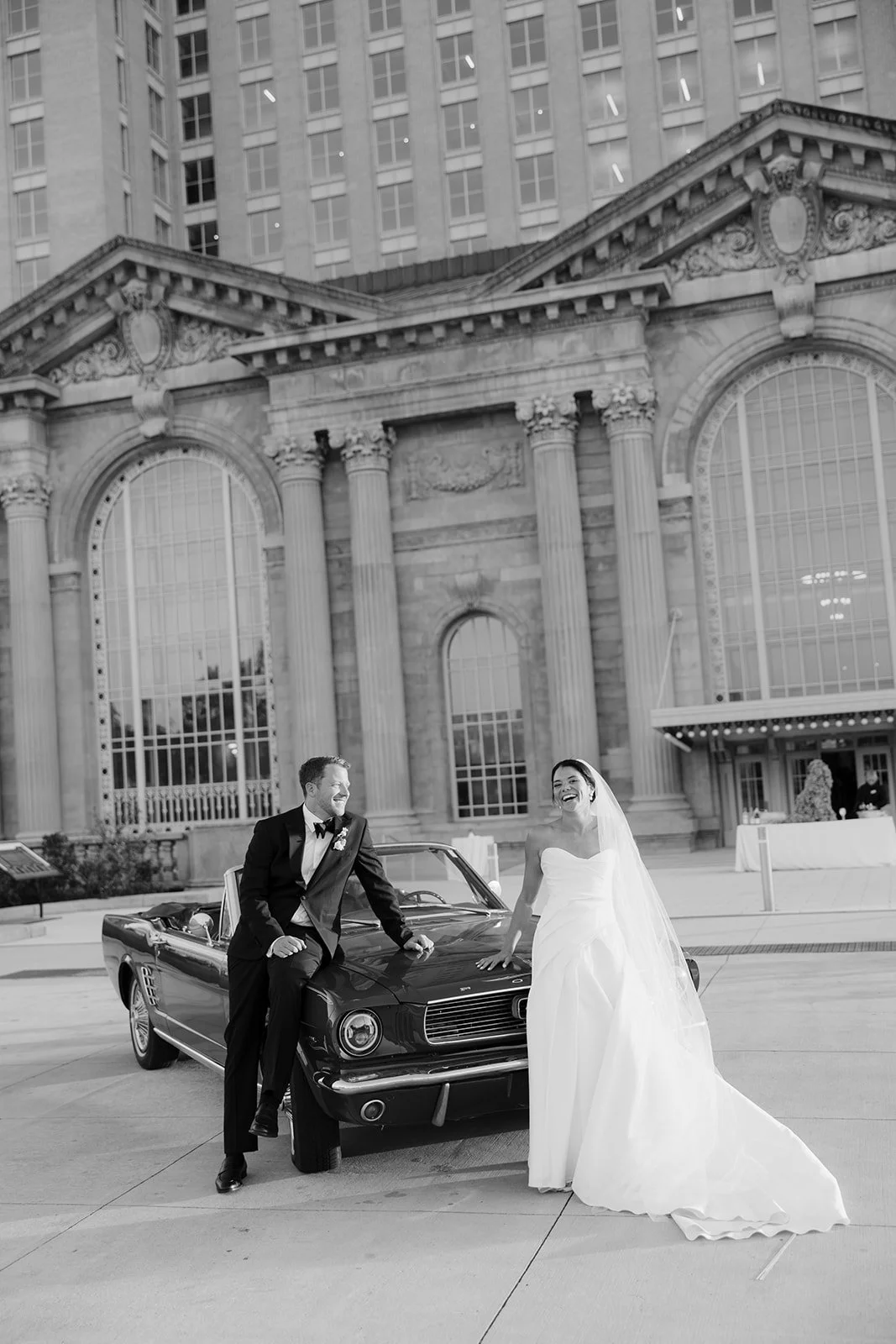 Bride and groom posing beside a vintage convertible outside an ornate historic building.
